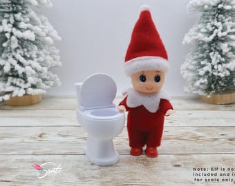 May include: A white miniature toilet with a closed lid. A small elf doll wearing a red and white Santa suit stands next to the toilet. Note: Elf is not included and is for scale only.