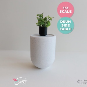 May include: A miniature white drum side table with a small potted plant. The plant has green leaves and is in a black pot. The table is labeled "1/6 SCALE" and "DRUM SIDE TABLE".