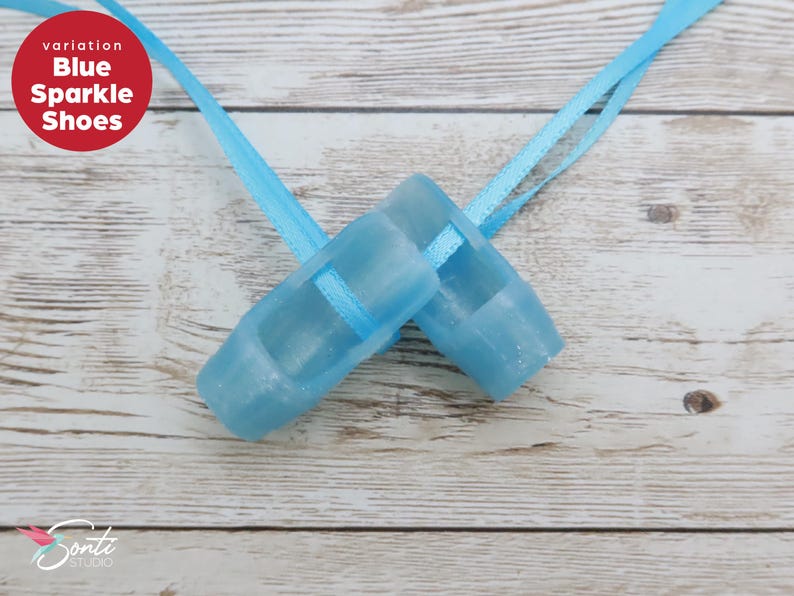 May include: Two light blue sparkle shoes with matching ribbons are displayed on a light wood surface. The shoes are small, possibly for a child or doll. The text "Blue Sparkle Shoes" is in a red circle.