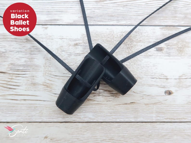 May include: A pair of black ballet shoes with long black ribbons, on a light wood grain surface. The shoes are a solid black colour and have a classic ballet slipper design. The text "variation Black Ballet Shoes" is in a red circle.
