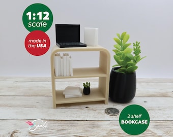 May include: A miniature 2-shelf bookcase in a light beige color, with a black laptop, white block, and stacks of white books. A small black potted plant and a larger plant in a black pot are also present. The image includes the text "1:12 scale" and "2 shelf BOOKCASE".