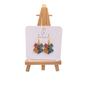 May include: A pair of gold-colored drop earrings featuring a colorful crest design with red, green, blue, and yellow enamel. The earrings are displayed on a white card with the text "Melanated Beauty Queens".