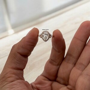Puede incluir: Un anillo de compromiso de diamante de forma pera con un engaste halo. El anillo está engastado en oro rosa y presenta diamantes más pequeños en la banda.
