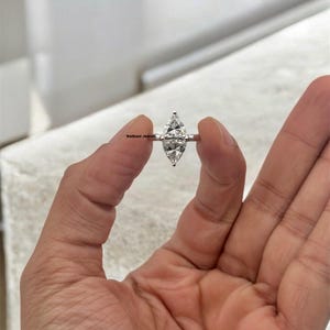 Puede incluir: Un anillo de plata con un diamante de forma marquesa. El diamante es claro y brillante. El anillo está en una mano.