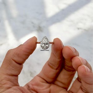 Puede incluir: Un anillo de compromiso de diamante de forma pera con engaste halo. El anillo está engastado en una banda de plata y presenta un solo diamante grande de forma pera rodeado de diamantes más pequeños.