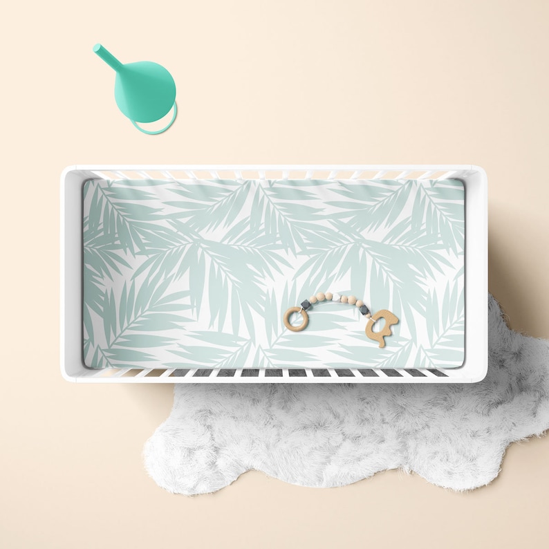 Op de afbeelding: Een wit babybedje met een lichtblauw en wit beddengoed met een palmbladmotief. Een houten baby speelgoed met een kralenketting ligt op het beddengoed.