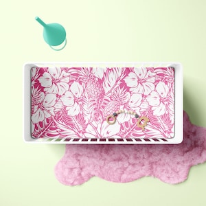 Puede incluir: Una cuna blanca con una sábana estampada con flores rosas y blancas. Una alfombra peluda rosa está en el suelo debajo de la cuna.