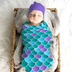 May include: A baby swaddled in a turquoise and purple mermaid scale swaddle blanket with a purple knit hat.