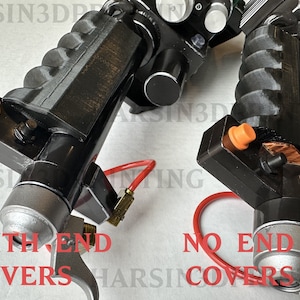 May include: Two black and silver 3D printed prop replicas of a fictional weapon. The replica on the left has a silver end cap, while the replica on the right has a raw, unfinished end. Both replicas have orange buttons and red tubing.
