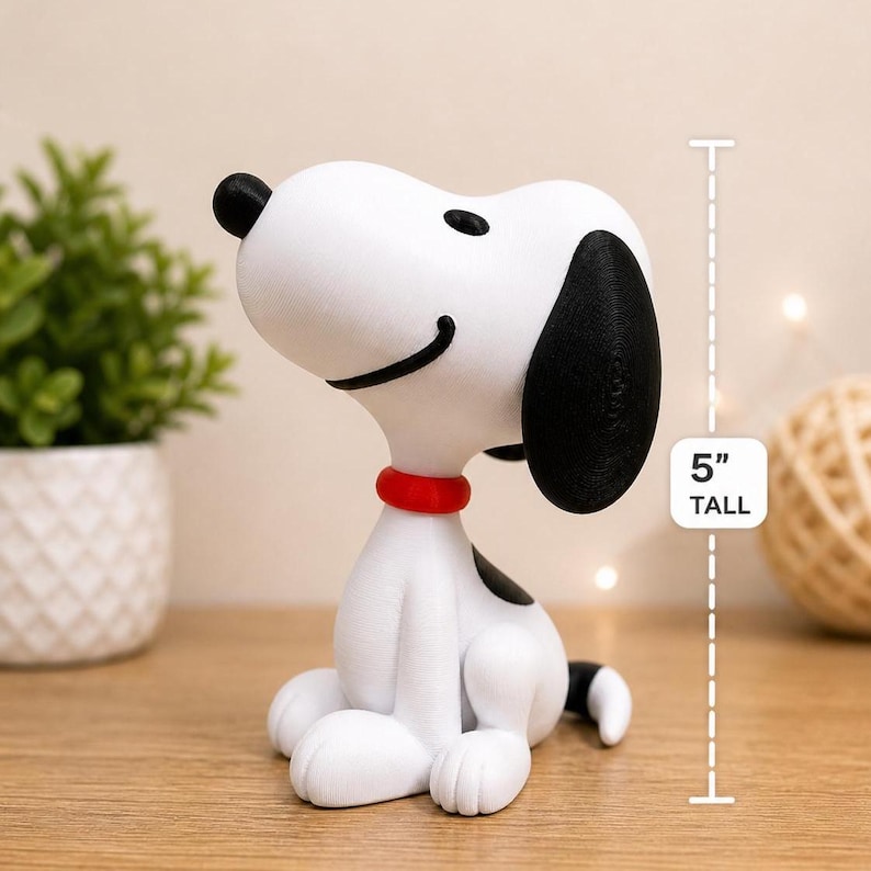 May include: A 5-inch tall Snoopy figurine. The white figure has black ears, a black spot on its back, and a red collar. Snoopy is sitting with a smiling expression. A small plant and a ball of twine are in the background.