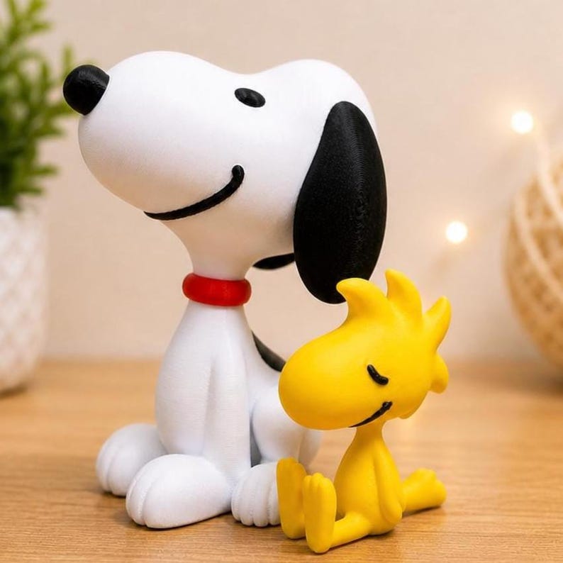 May include: A white Snoopy figurine with black ears and a red collar sits next to a yellow Woodstock figurine. The figures are on a wooden surface. The Snoopy figurine has a black spot on its back.
