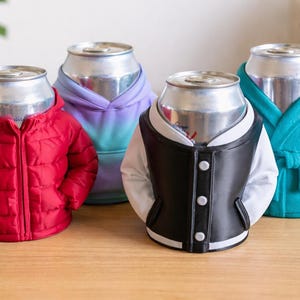 Puede incluir: Cuatro latas de bebidas vestidas con trajes en miniatura. Una lleva una chaqueta acolchada roja, otra una sudadera con capucha morada y azul, una tercera una chaqueta universitaria en blanco y negro y la última una bata de baño turquesa. Las latas están sobre una superficie de madera.