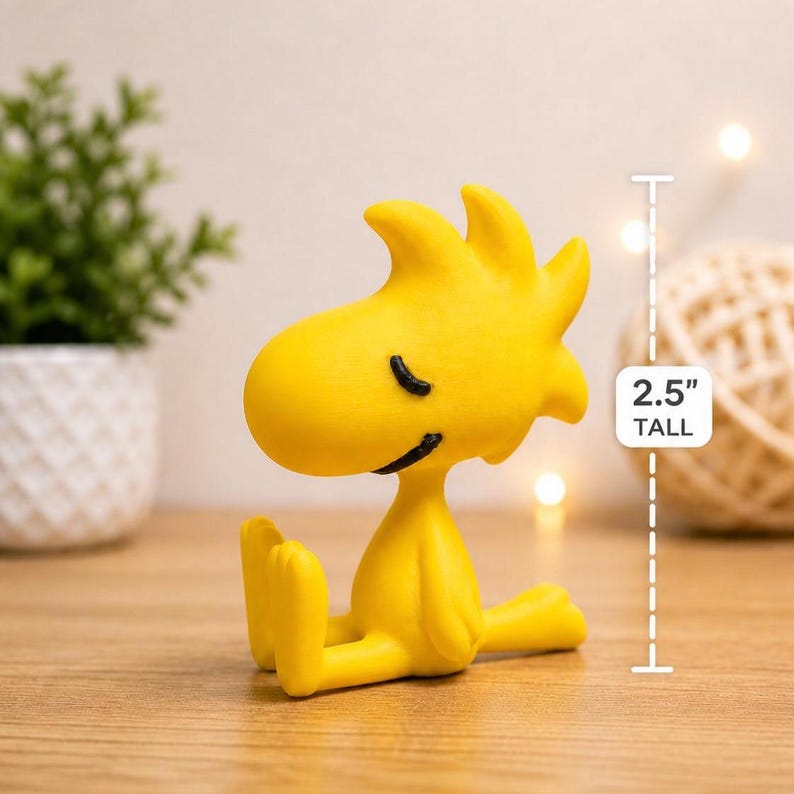 May include: A yellow figurine of Woodstock from the Peanuts comic strip, sitting with his legs extended. The figurine is 2.5 inches tall and has black eyes and a smiling beak. The figure is on a wooden surface.