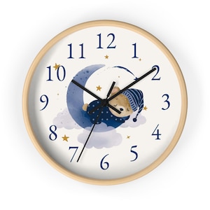 May include: A white clock with a blue crescent moon and a sleeping bear wearing a blue and white hat. The clock face has black hands and blue numbers. The moon has gold stars on it.