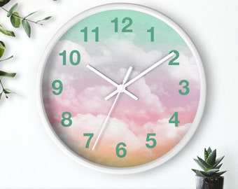 May include: A white-framed wall clock with a pastel sky design. The clock face features a gradient of colors, including pink, orange, and green, with white clock hands and green numerals. The clock is a decorative item.