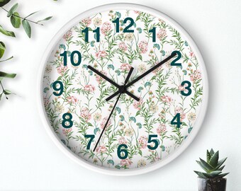 May include: White wall clock with a floral pattern featuring pink, blue, and yellow flowers on a white background. The clock has black hands and dark teal numbers. A small potted plant is visible in the lower right corner.