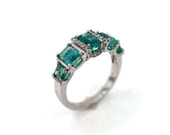 May include: A silver ring featuring five rectangular, emerald-cut, teal-colored gemstones. The center stone is accented with small, clear gemstones. The ring is set against a white background, highlighting the jewelry's design.