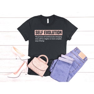 Puede incluir: Camiseta negra con el texto "SELF EVOLUTION" en un recuadro rosa. El texto debajo del recuadro dice "The process of continuously changing ones self to a higher or more complex way of being."