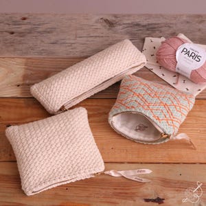 Puede incluir: Tres estuches hechos a mano con cremallera en crema y verde menta con bordados naranjas. Un estuche es rectangular, otro cuadrado y el tercero tiene forma larga y estrecha. También se ve una madeja de hilo rosa y una muestra de tela.