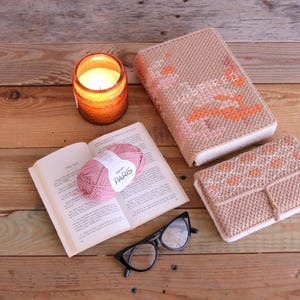May include: A still-life arrangement featuring a lit orange candle in a glass jar, an open book with a pink yarn ball labeled "PARIS", and black cat-eye glasses. Two beige crocheted book covers with orange and pink accents are also present.