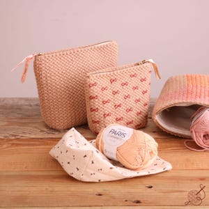May include: A collection of handmade, peach-colored knitted pouches and a woven basket with a zipper. One pouch has a bow pattern. A ball of peach-colored yarn and a patterned fabric are also present. The word "PARIS" is visible on the yarn label.