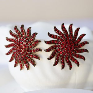 May include: A pair of red rhinestone sunburst earrings with a gold tone finish.
