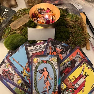 Puede incluir: Una colección de cartas del tarot, incluyendo la carta "El Mundo", dispuestas sobre una mesa con musgo, una vela encendida en un cuenco dorado y otros objetos místicos. Las cartas presentan ilustraciones coloridas y texto, creando un tema espiritual y esotérico.