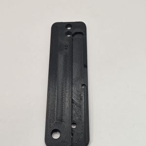Può includere: Un oggetto nero rettangolare stampato in 3D con una serie di fori e un design scanalato. L'oggetto sembra essere un componente o parte di un meccanismo più grande. La superficie ha un aspetto strutturato.