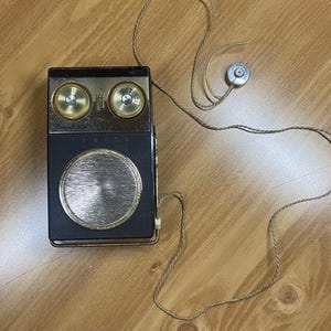 May include: A black vintage transistor radio with a gold speaker grille and two gold knobs. The radio has a cord with a small earpiece attached.
