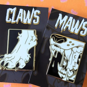 May include: Two enamel pins on black cards. One pin features a white claw design with gold trim, and the word "CLAWS" in a bold font. The other pin shows a white maw design with gold trim, and the word "MAWS".