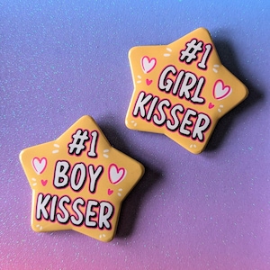 May include: Two yellow star-shaped badges with pink hearts and black text. One badge says "#1 GIRL KISSER" and the other says "#1 BOY KISSER".