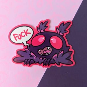 Op de afbeelding: Een sticker met een cartoonmonster met grote roze ogen en een tekstballon met de tekst "Fuck". Het monster is paars met roze accenten en een brede, tandige grijns.