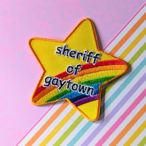 Può includere: Toppa gialla a forma di stella con la scritta "sheriff of gaytown". La toppa presenta un design arcobaleno ed è posta su uno sfondo rosa e a righe. È un articolo tessile.