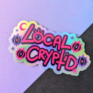 Può includere: Un adesivo olografico con le parole "Local Cryptid" in grassetto, rosa, bordate di nero. L'adesivo ha un bordo frastagliato e presenta elementi decorativi come stelle, lune e forme stilizzate a occhio in viola e rosa.