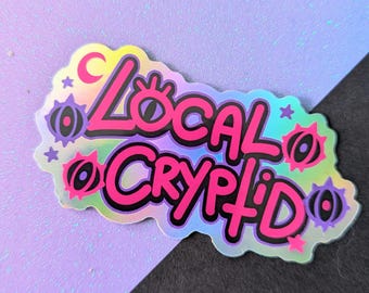 Könnte beinhalten: Ein holografischer Aufkleber mit den Worten "Local Cryptid" in einer kräftigen, pinkfarbenen Schrift, die schwarz umrandet ist. Der Aufkleber hat einen gezackten Rand und enthält dekorative Elemente wie Sterne, Monde und stilisierte Augenformen in Lila und Pink.