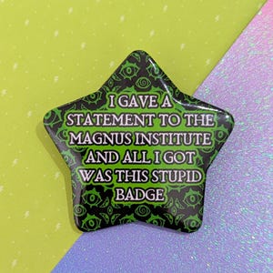 Puede incluir: Una insignia en forma de estrella con un diseño verde y negro. La insignia tiene el texto blanco "I GAVE A STATEMENT TO THE MAGNUS INSTITUTE AND ALL I GOT WAS THIS STUPID BADGE". El fondo es una mezcla de verde, morado y azul.