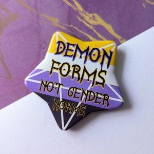 Puede incluir: Un botón en forma de estrella amarillo, morado y negro con el texto "Demon Forms Not Gender Norms" en negro.