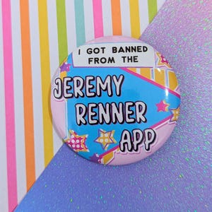 Könnte beinhalten: Ein runder, rosa Button mit dem Text "I GOT BANNED FROM THE JEREMY RENNER APP". Der Button hat ein blaues und gelbes Design mit Sternakzenten. Der Hintergrund ist eine bunte Mischung aus Streifen und Glitzer.