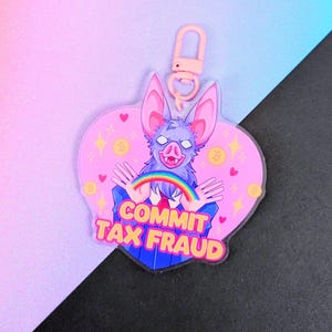 May include: A heart-shaped acrylic keyring featuring a purple bat character with large ears and white eyes. The bat holds a rainbow arch. The text COMMIT TAX FRAUD is printed in yellow and orange. It includes a light pink plastic lobster clasp.