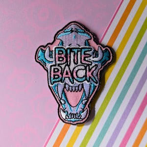 May include: Embroidered patch with a skull design and the text "BITE BACK". The patch uses teal, pink, and white thread, with a black border. It's set against a pink backdrop with rainbow stripes.
