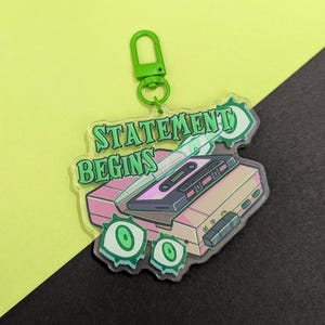 May include: A clear acrylic keychain with a green clasp. The keychain features a pink and tan cassette player with a cassette tape, green eye-shaped designs, and the words "STATEMENT BEGINS" in green lettering.