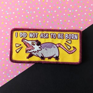 Peut inclure: Un patch brodé jaune avec une bordure rouge représentant un opossum de dessin animé avec le texte "Je n'ai pas demandé à naître."