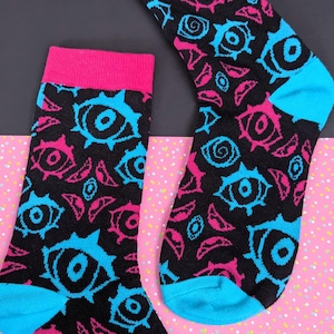 May include: A pair of black crew socks featuring a repeating pattern of stylized eyes in vibrant hot pink and bright cyan. The socks have solid hot pink cuffs at the top and solid bright cyan heels and toes, creating a bold contrast against the black background.