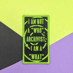 May include: A rectangular embroidered patch with a black background and a bright lime green border. The text reads I AM NOT A "WHO" ARCHIVIST; I AM A "WHAT" in lime green capital letters, surrounded by a circular graphic design element.