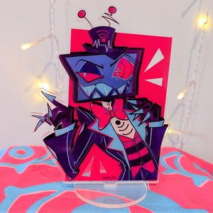 Może przedstawiać: Kolorowy stojak akrylowy przedstawiający postać z kreskówki z głową w kształcie telewizora, ubraną w garnitur i muszkę. Postać ma groźny wyraz twarzy z czerwonym okiem i ostrymi zębami. Tło jest w żywym różu.