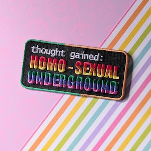 Op de afbeelding: Geborduurde patch met de tekst "thought gained: HOMO-SEXUAL UNDERGROUND" in regenboogkleuren op een zwarte achtergrond. De patch heeft een groene rand en staat op een roze en regenboog gestreepte achtergrond.