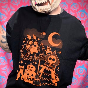 Puede incluir: Camiseta negra con un diseño naranja de Halloween. El diseño incluye una polilla, una bruja, una calabaza, murciélagos y estrellas. La camiseta es de cuello redondo y tiene mangas cortas.
