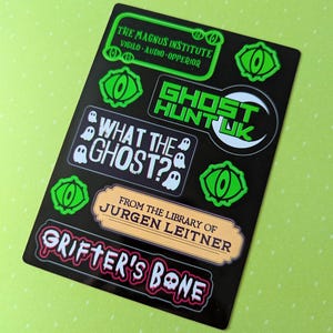 May include: A black sticker sheet with various text and graphic designs. The designs include phrases like "GHOST HUNT UK" and "GRIFTER'S BONE", along with eye and ghost illustrations. The text "THE MAGNUS INSTITUTE" is also present.
