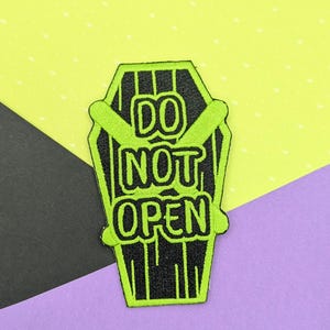 May include: An embroidered patch shaped like a coffin with a black background and bright lime green borders and text. The text reads DO NOT OPEN in a bold, rounded font. Two lime green bars are crossed horizontally over the centre of the coffin.