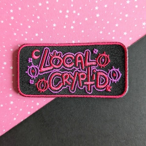 Op de afbeelding: Een zwarte geborduurde patch met roze en paarse stiksels. De patch zegt "Local Cryptid" met een halve maan en gestileerde ogen.